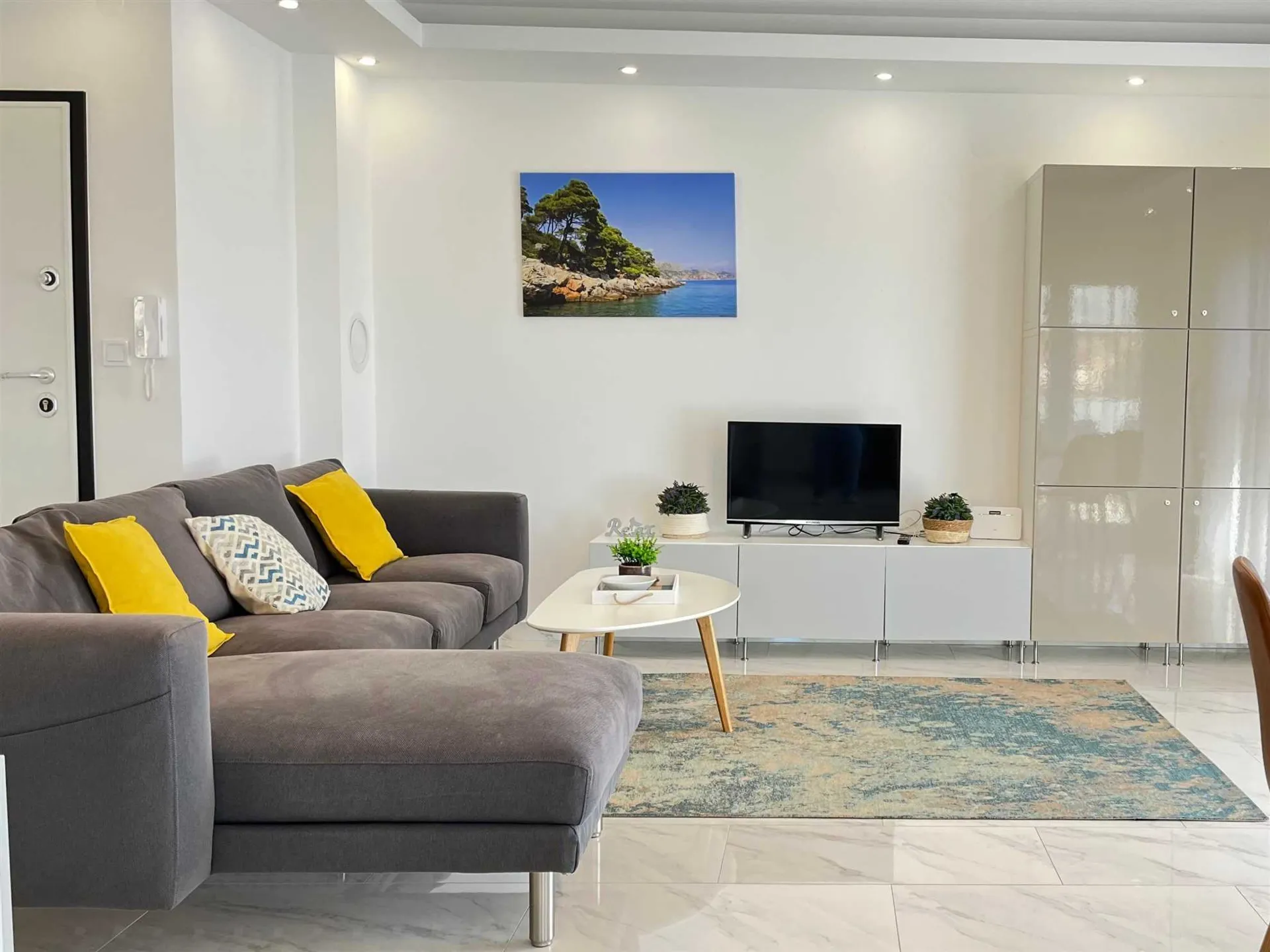 A contemporary living room with a gray sectional sofa adorned with yellow cushions, a white coffee table, glossy white cabinets, and a picture of a coastal landscape on the wall in Apartment Buby